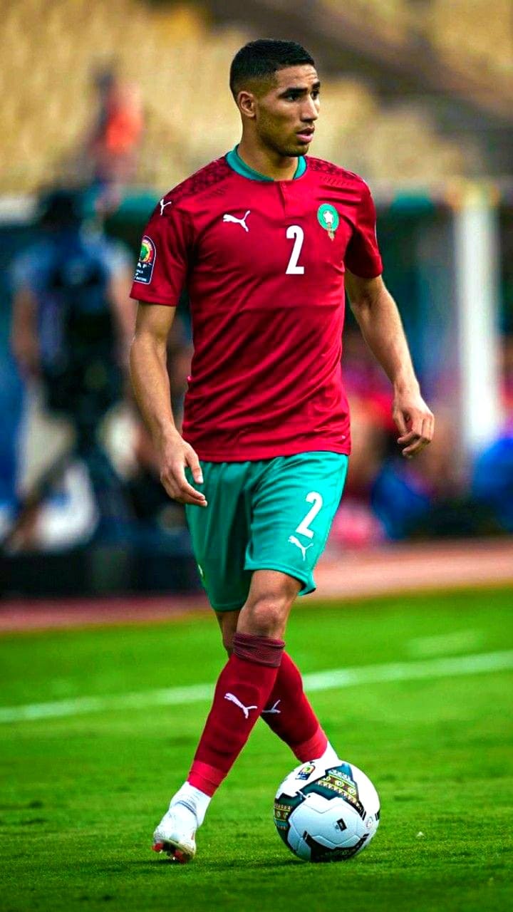 A soccer player in a red and green Morocco jersey with the number 2 is dribbling a ball on a field during a match, as fans around the world watch live through Nordic IPTV.